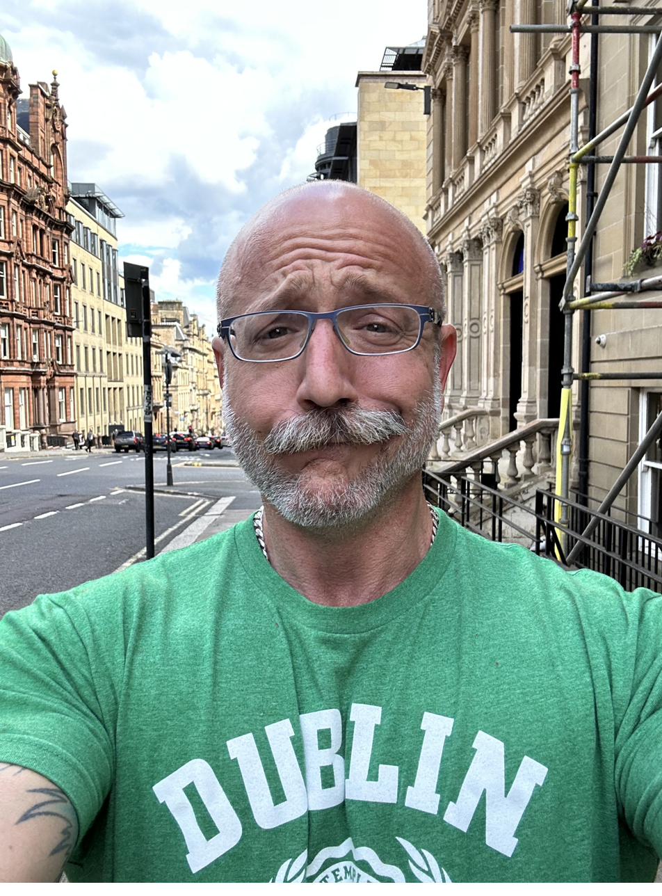 A selfie of Barclay Barrios walking along a street lined with classical-looking buildings.